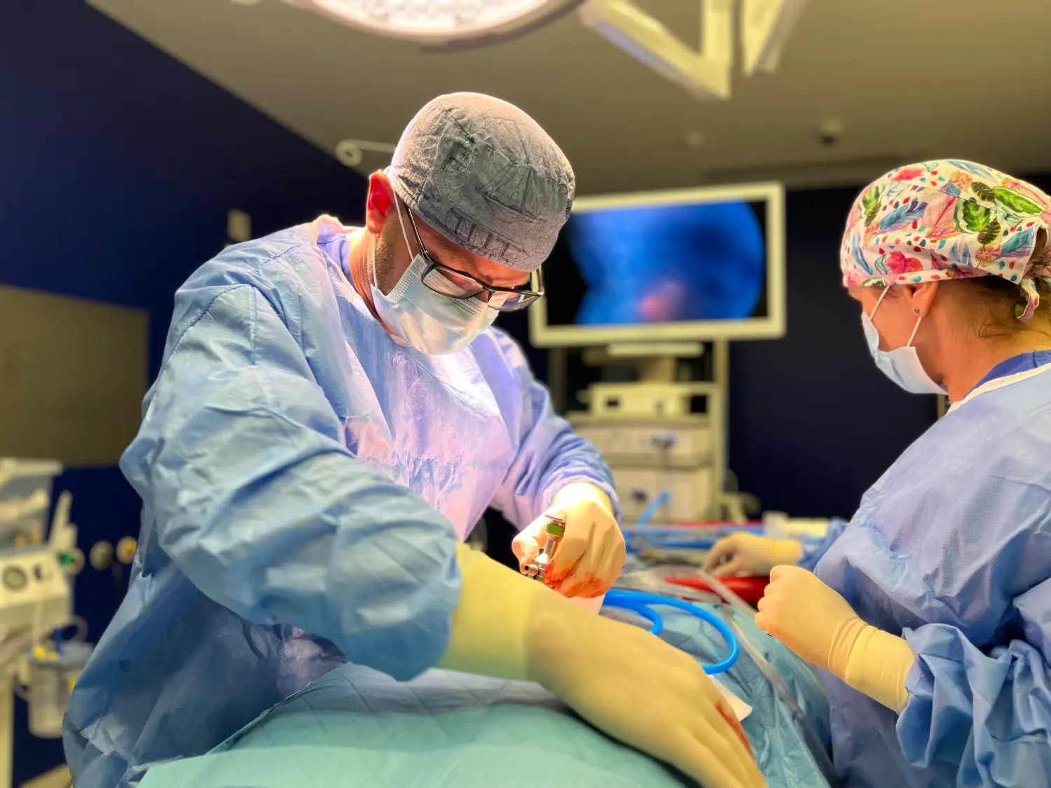 Dwóch chirurgów w fartuchach i maskach przeprowadza operację w jasno oświetlonej sali operacyjnej.