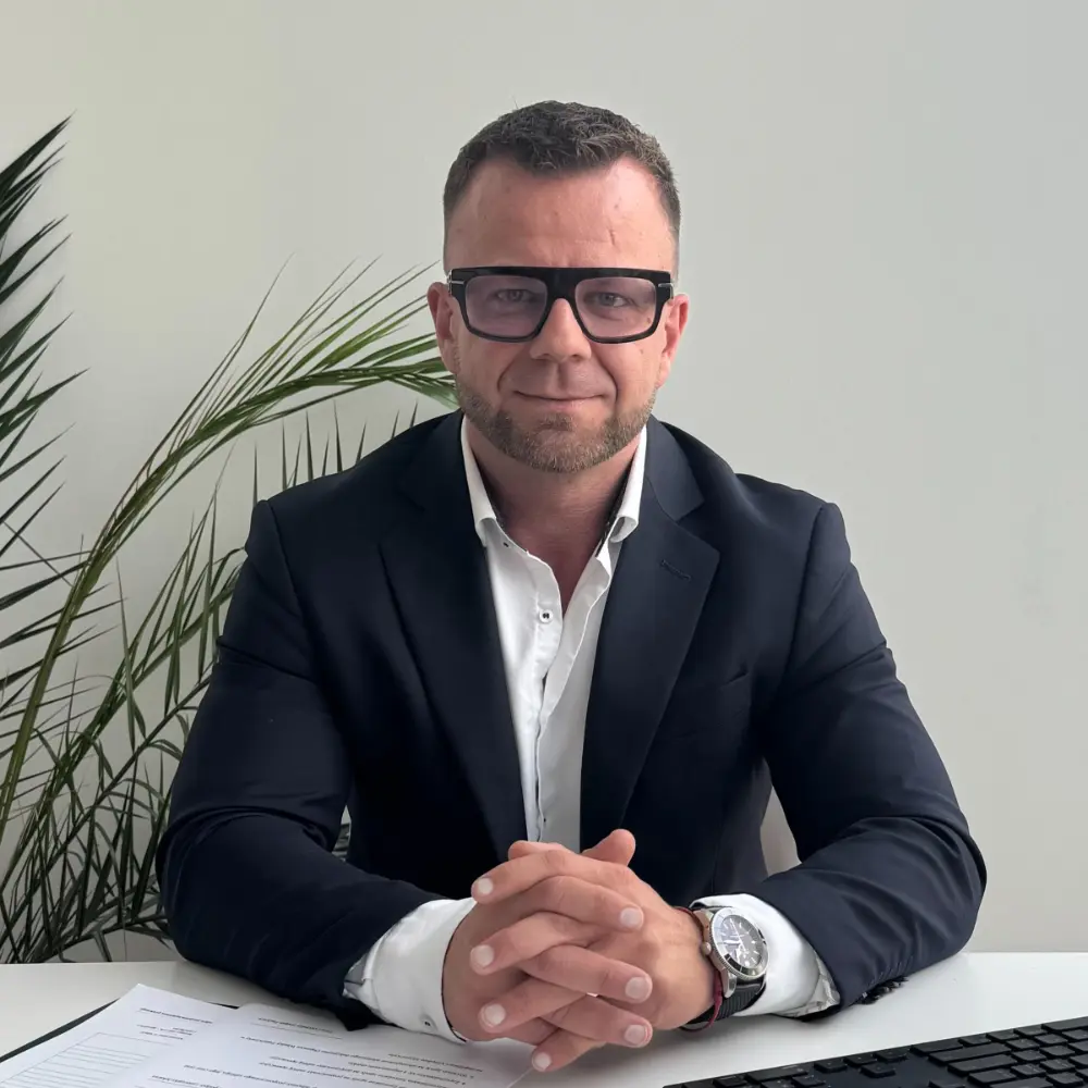 Mężczyzna w garniturze i okularach siedzi przy biurku z papierami, w tle roślina, ręce złożone.