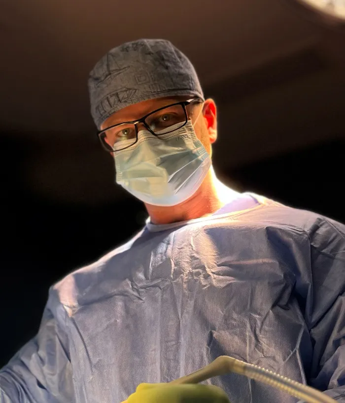 Chirurg w masce, okularach i fartuchu patrzy w dół, trzymając narzędzia chirurgiczne w jasnym świetle.