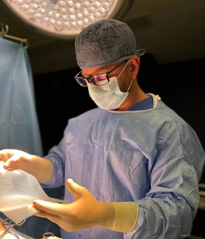 Chirurg w fartuchu i rękawiczkach bada siatkę chirurgiczną w świetle sali operacyjnej.